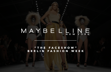 Image of a dynamic fashion runway scene at Berlin Fashion Week featuring the Maybelline Make-Up Runway. The runway is illuminated by dramatic lighting with models showcasing bold, artistic makeup looks. The audience of fashion VIPs is visibly engaged, capturing the event on their devices. The backdrop features large, vibrant projections enhancing the atmospheric mood of the show.